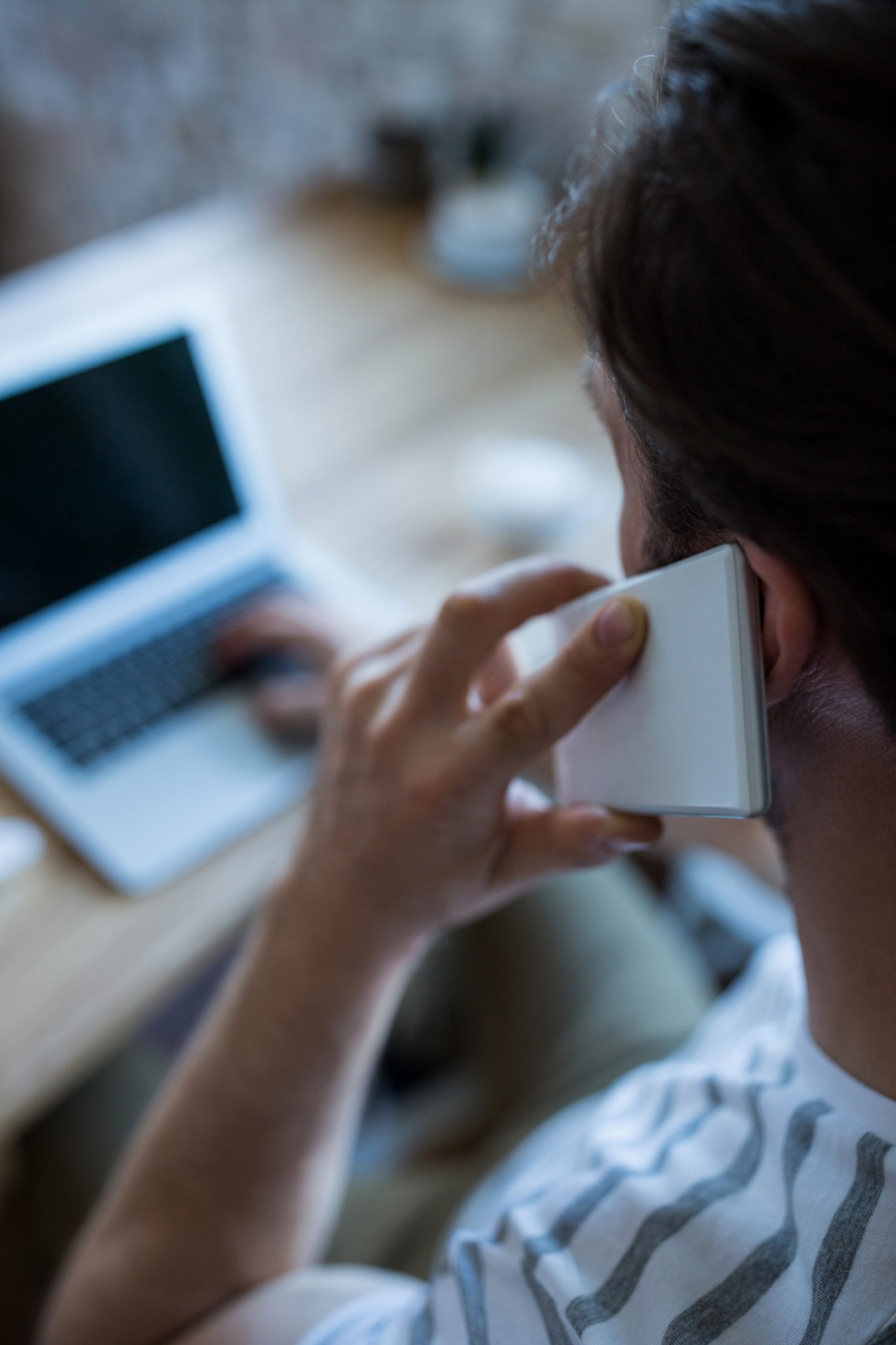 Rear view of man talking on the mobile phone and using laptop in coffee shop